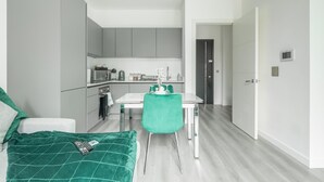 Une chambre moderne et compacte, avec une couvre-lit verte, une petite table et une chaise. À côté se trouve une kitchenette équipée d’un micro-ondes, d’une bouilloire et de placards.