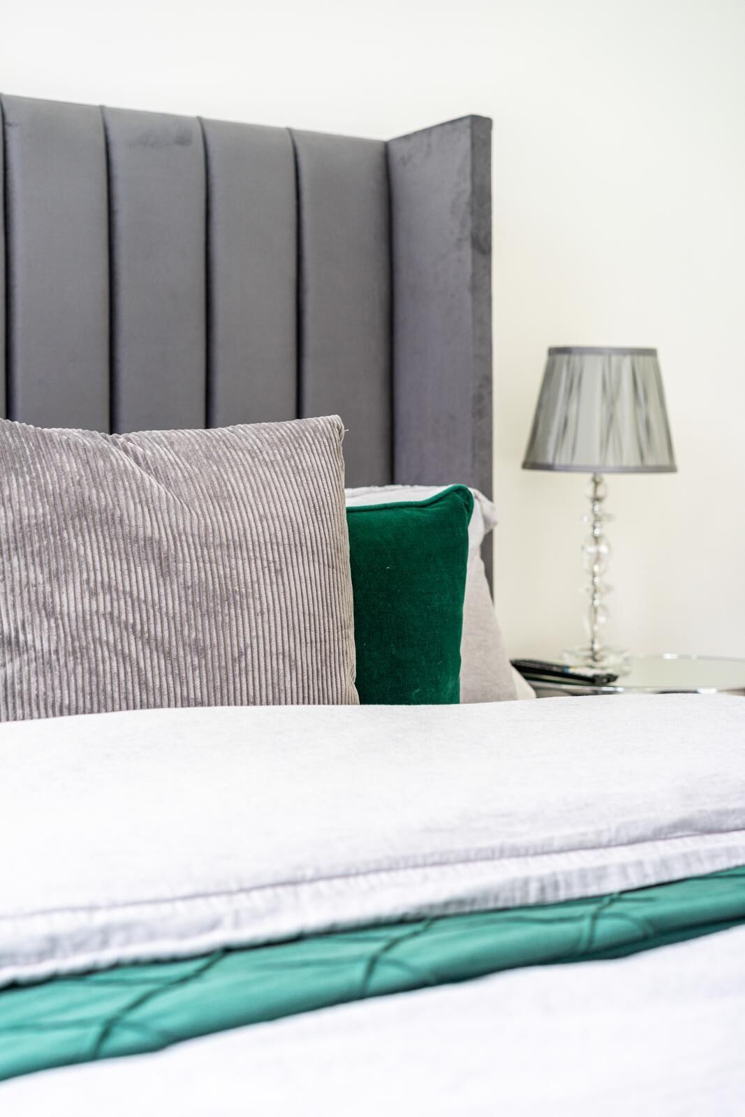 Un lit avec une tête de lit grise, un oreiller gris et un oreiller vert. Une lampe se trouve sur une table de chevet.