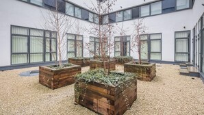 Une cour intérieure agrémentée de jardinières en bois où sont plantés des arbres et des arbustes, le tout entouré d’un bâtiment doté de grandes fenêtres et d’une façade blanche.