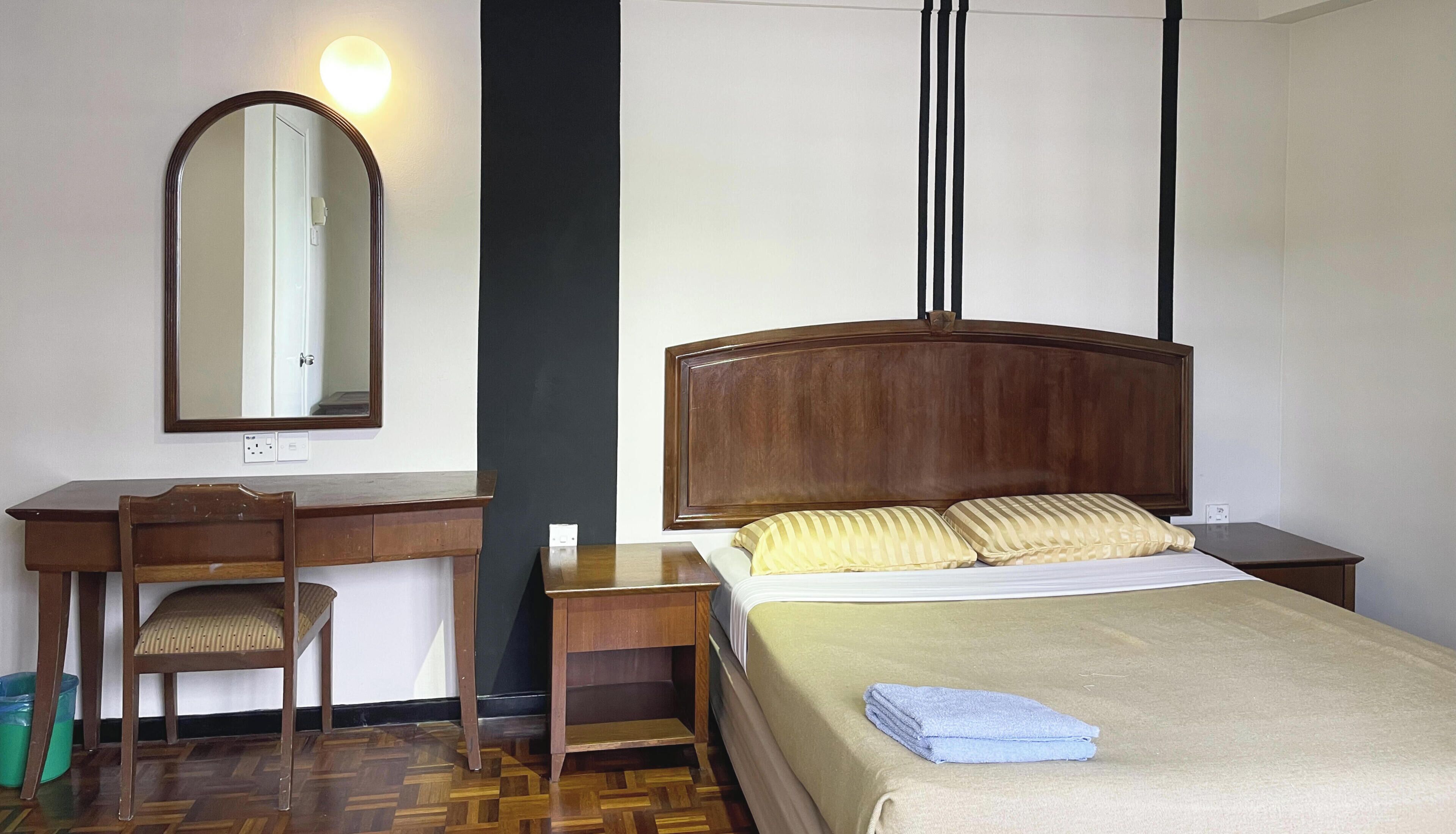 A hotel room with a wooden desk, a chair, a bed with a wooden headboard, a nightstand, and a mirror on the wall.