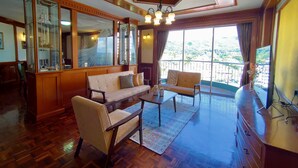 Un salón amplio con sofá, sillones, una mesa de centro y una televisión. La habitación cuenta con suelo de madera, un ventanal con vistas y un techo con candelabro.