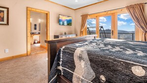 Un dormitorio con una cama grande, cabecera de madera, ventilador de techo, un cuadro, un baño con inodoro y lavamanos, y vistas a montañas a través de amplios ventanales.