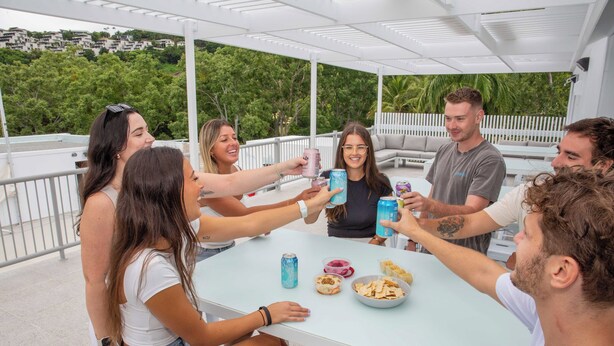 A group of people toasting with drinks around a table with snacks and a dessert bowl, under a white pergola with a view of greenery and buildings. - Bounce Airlie Beach (Airlie Beach)