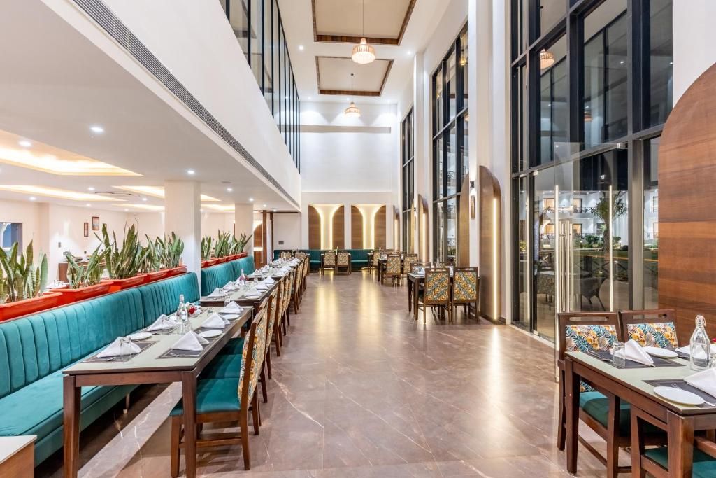 A spacious hotel dining area with long tables and booths, large windows, and indoor plants.