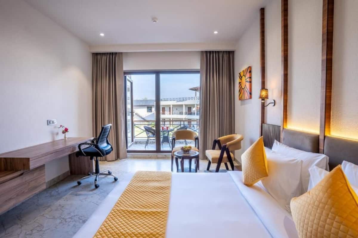 A hotel room with a bed, desk, and chairs. There is a view of a balcony and buildings outside.