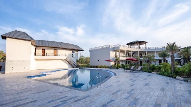 A pool area with a clear blue pool, a covered seating area with umbrellas, and a building with a balcony. - Shervani Pebbles And Pine, Corbett (Ramnagar)