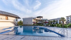 A swimming pool with a modern design, surrounded by a landscaped area with palm trees and a building with balconies. - Shervani Pebbles And Pine, Corbett (Ramnagar)