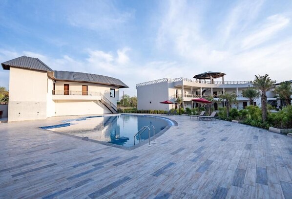 A pool area with a clear blue pool, a covered seating area with umbrellas, and a building with a balcony. - Shervani Pebbles And Pine, Corbett (Ramnagar)