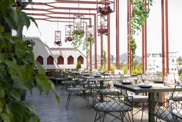 An outdoor dining area with tables and chairs, hanging plants, and a view of buildings in the background. - Hotel Bawa Udaipur (Udaipur)