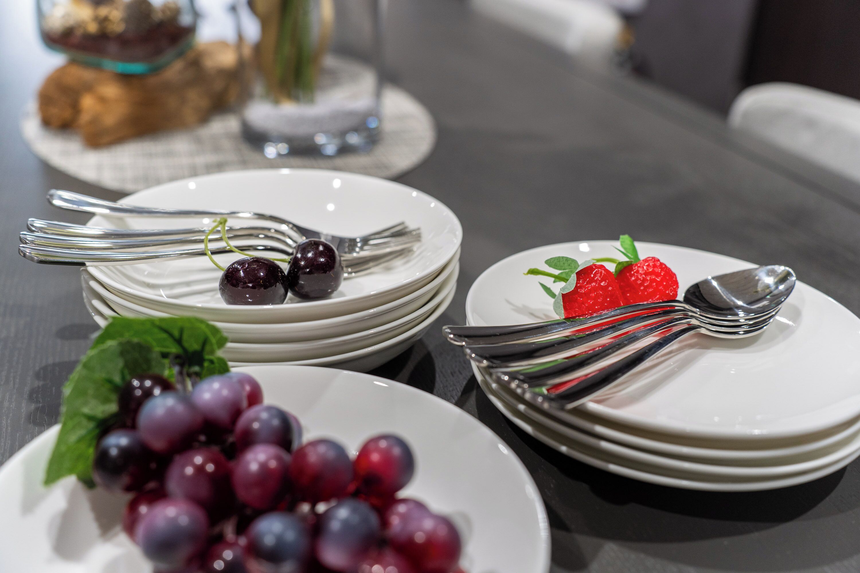 A table set with white plates, silverware, and a variety of fruits including grapes and strawberries.