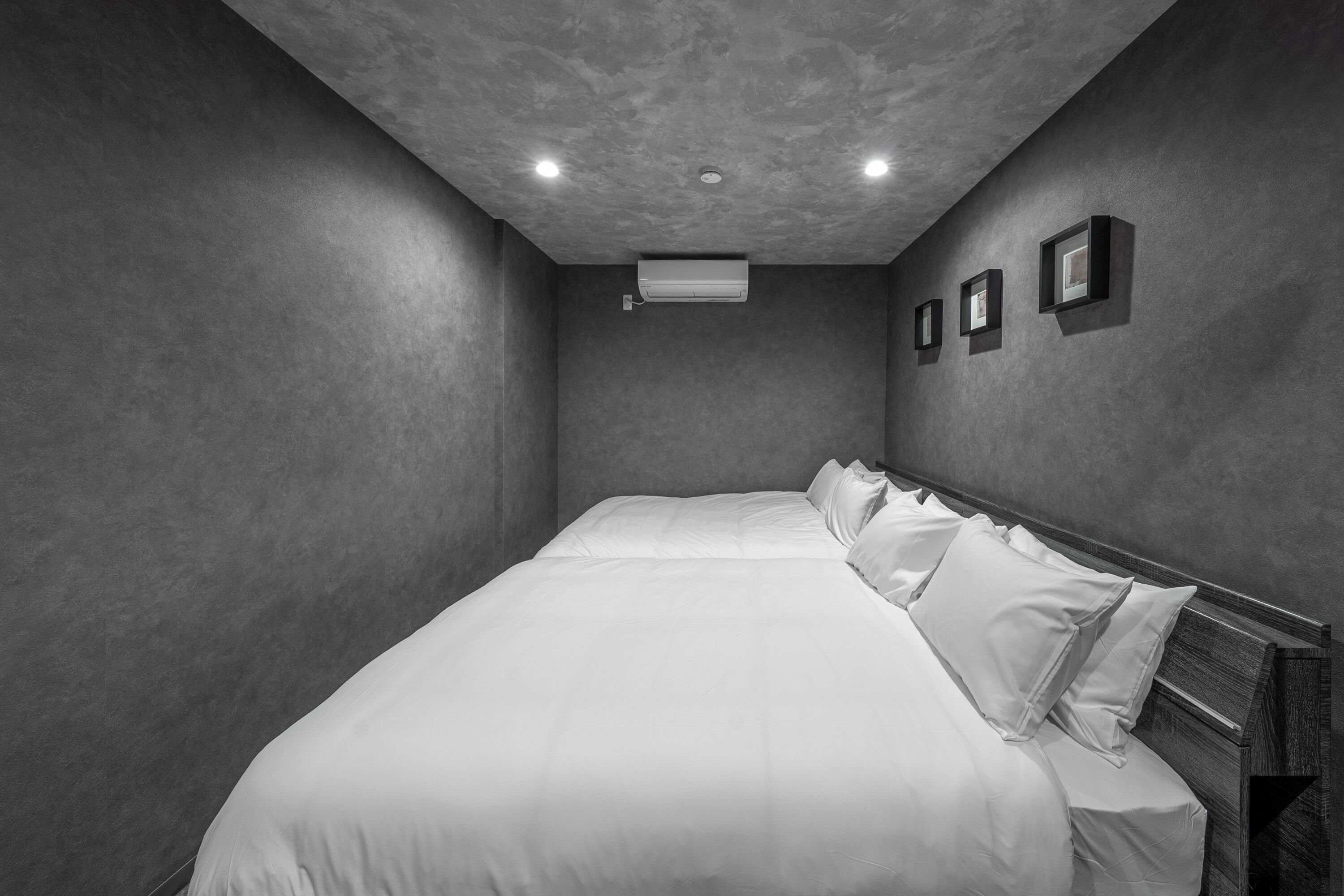 A monochrome hotel room with a large bed, a ceiling air conditioner, and three framed pictures on the wall.
