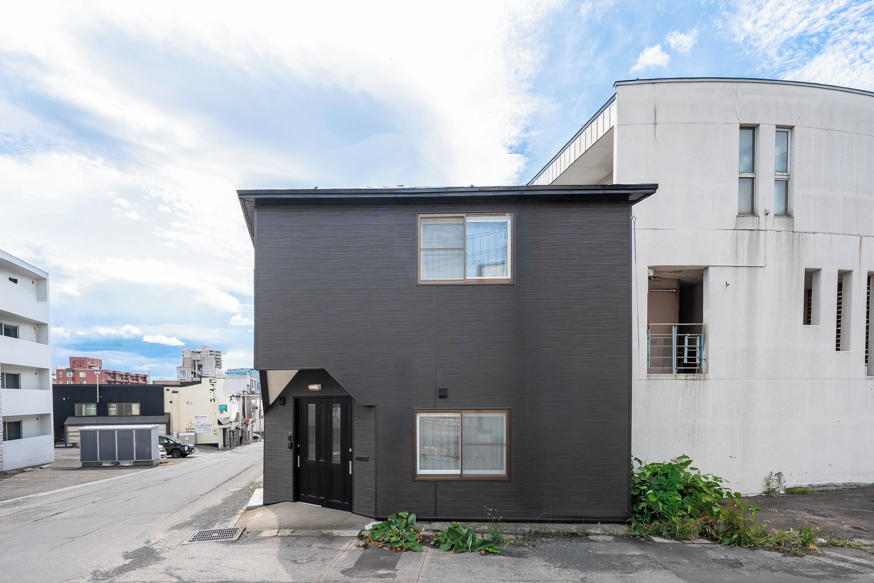 A modern two-story building with a dark facade, a small entrance, and a window on the ground floor.