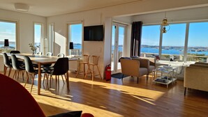 A spacious living area with a dining table, chairs, a sofa, and a TV mounted on the wall. Large windows provide a view of the sea. - Panoramic view on Ellös top with 500m to sea | SE09125 (Ellös)