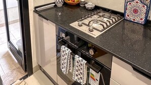 Eine Küche mit schwarzem Arbeitsplatz, einem Gasherd und einer Mikrowelle. Auf der Arbeitsfläche befinden sich dekorative Elemente, darunter eine verzierte Vase, eine Obstschale und ein Gewürzbehälter. Im Hintergrund ist ein Fenster zu sehen.