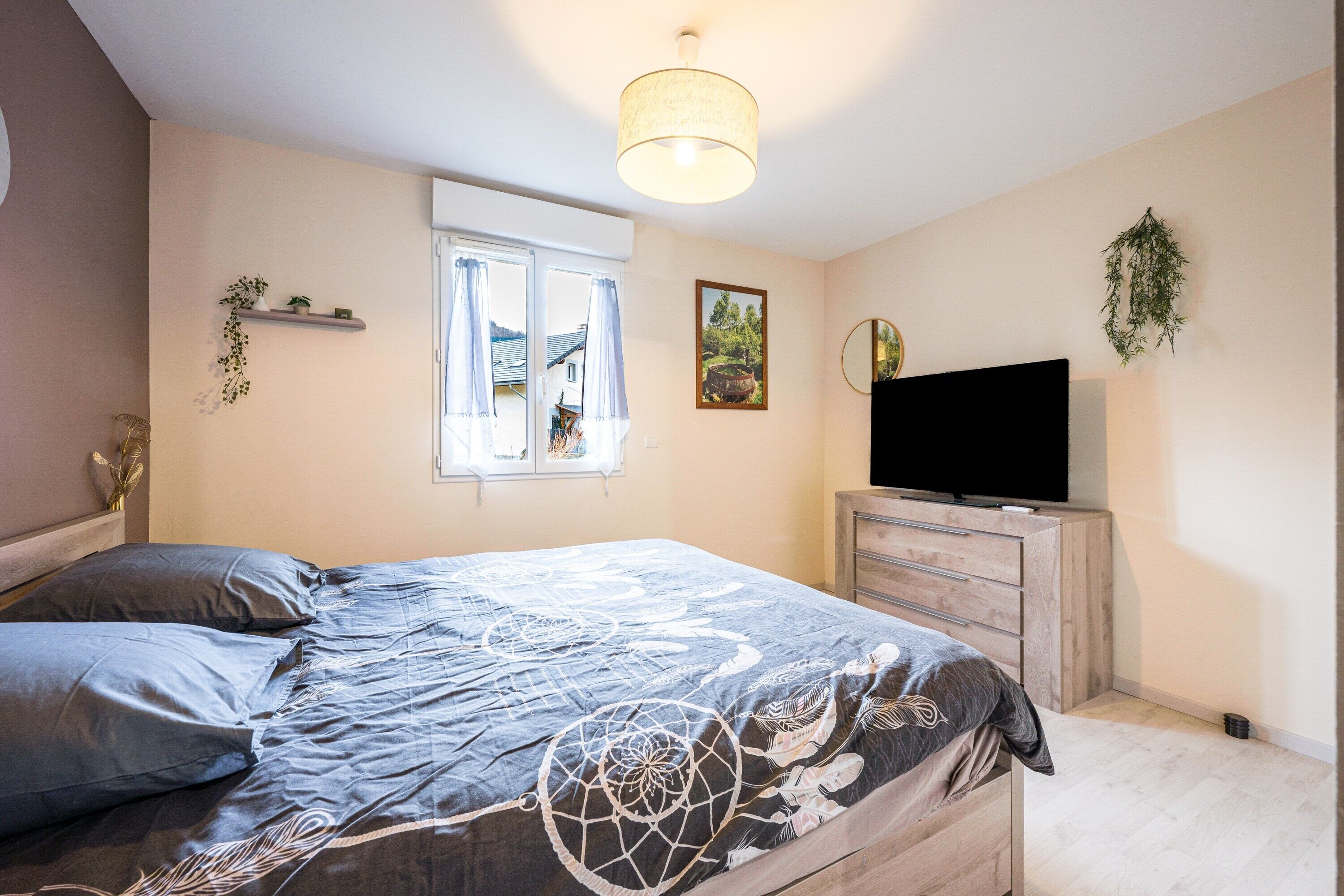 Een slaapkamer met een bed, een televisie, een raam met gordijnen, een wandplank met planten en een schilderij.
