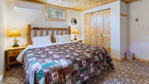 Un dormitorio con techo de madera, una cama con colcha estampada, dos mesitas de noche con lámparas, un ventanal con persianas y un armario de madera.