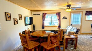 Una habitación acogedora con techo de madera, una mesa redonda para comer rodeada de sillas de madera, un televisor montado en la pared y una ventana con cortinas.