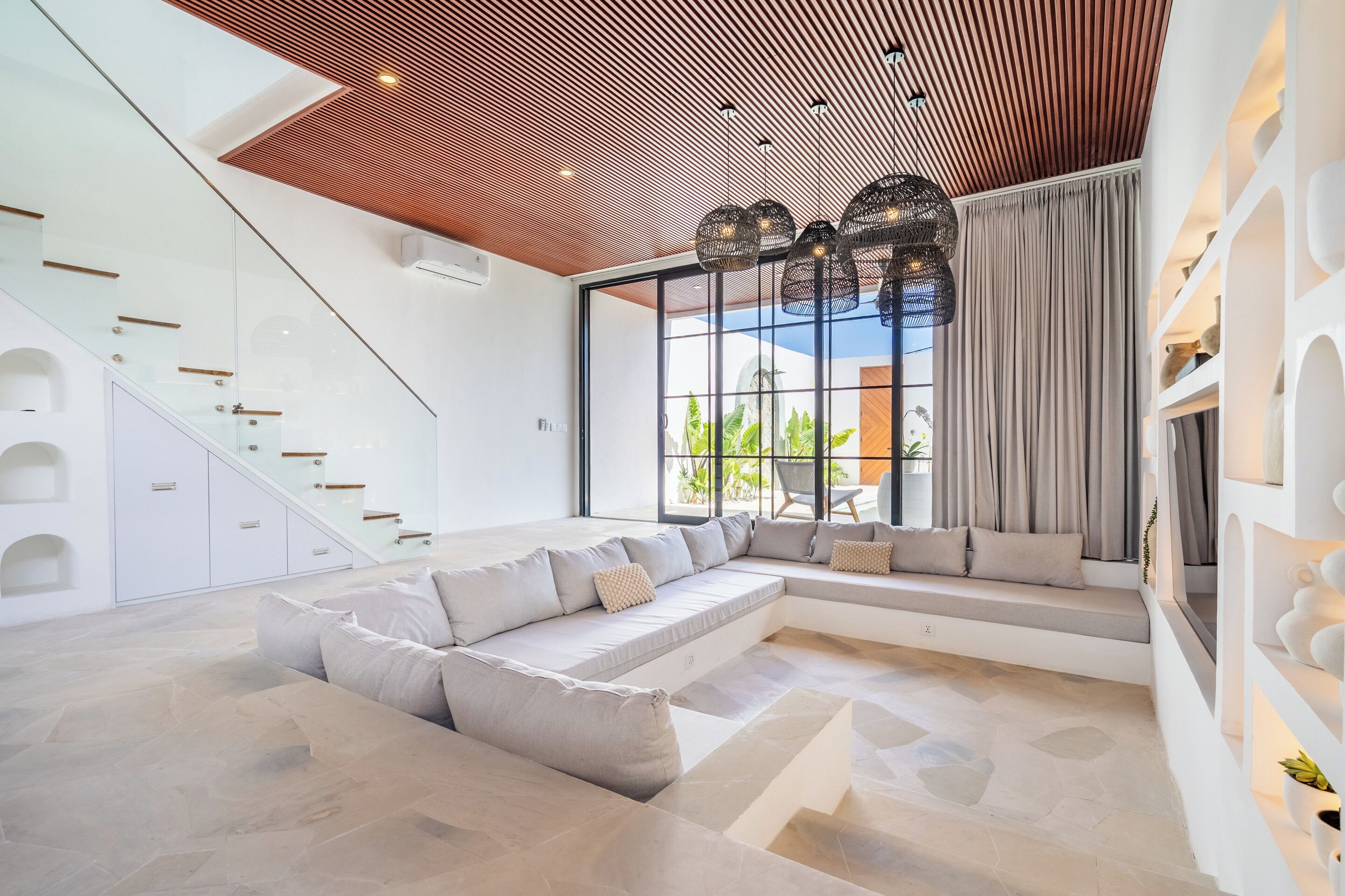 Een moderne woonkamer met een hoekbank, een trap met glazen balustrades en een groot raam met gordijnen.