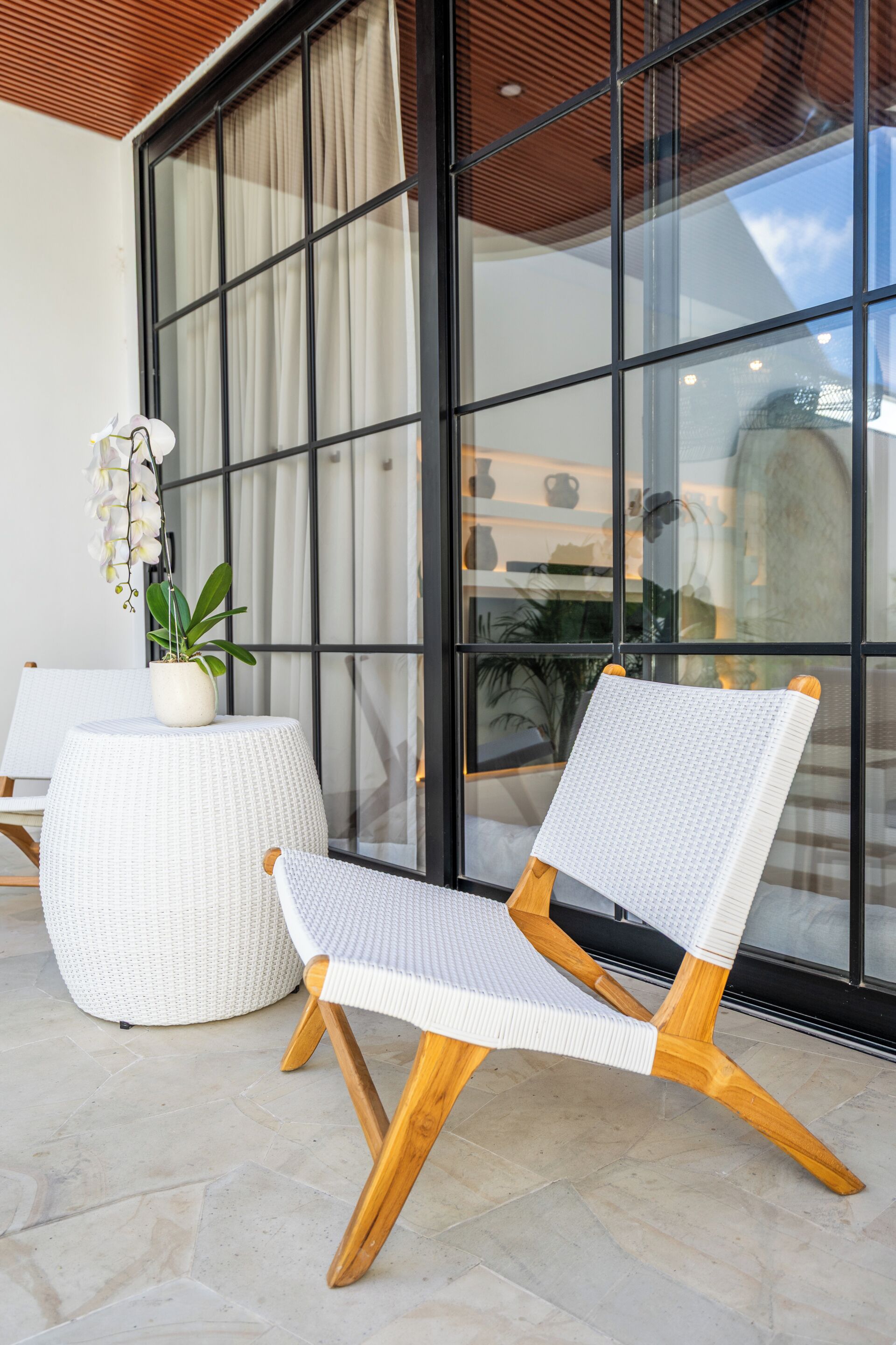 Une chaise en osier avec des pieds en bois et un siège tressé, placée sur une terrasse carrelée agrémentée d’une plante en pot et d’une table ronde.