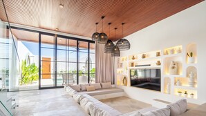 A modern living room with a large sectional sofa, a TV mounted on the wall, and a ceiling with wooden slats. - Villa Nuusa Bingin (Pecatu)