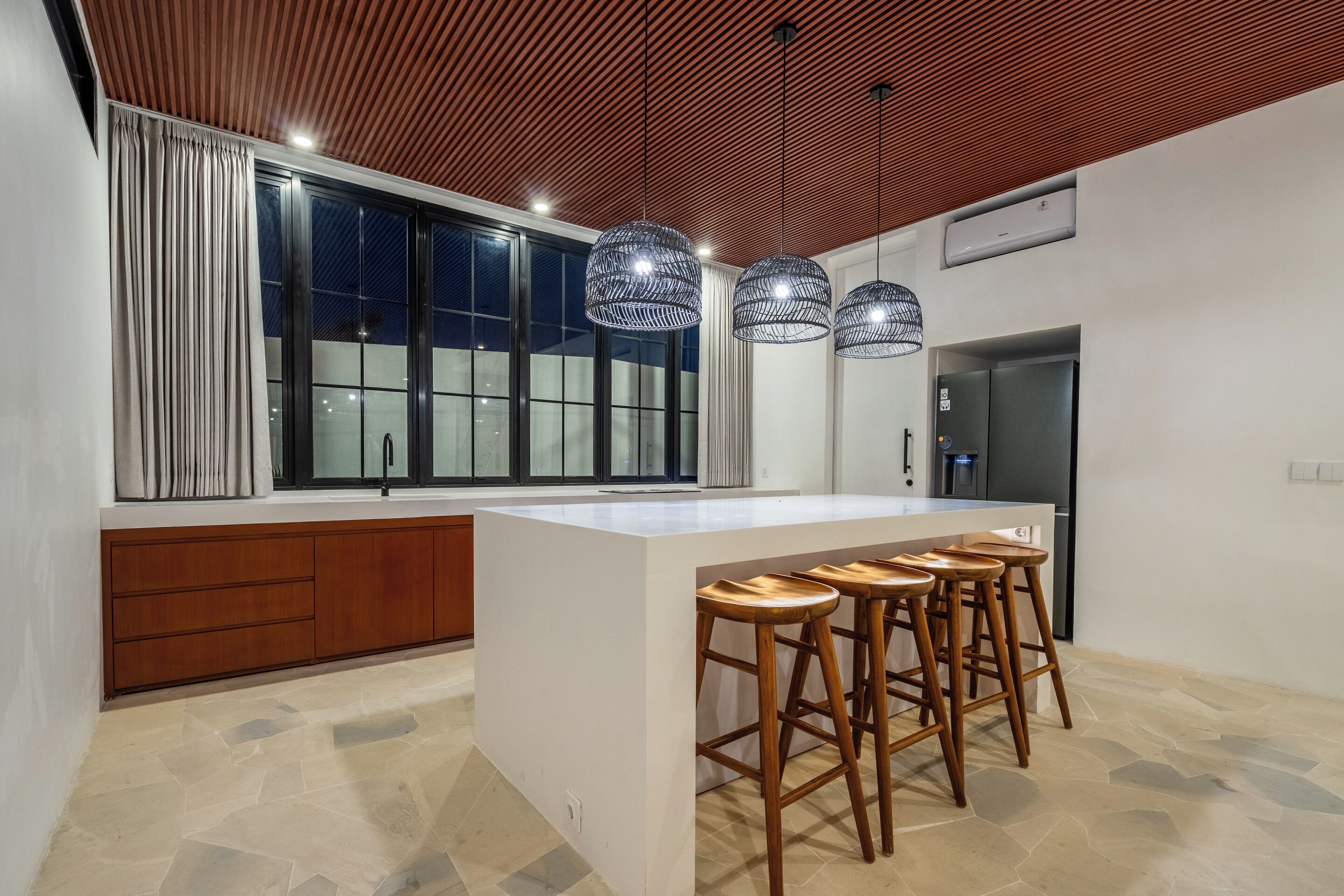 A modern kitchen with a large island, wooden bar stools, pendant lights, and a view through large windows.