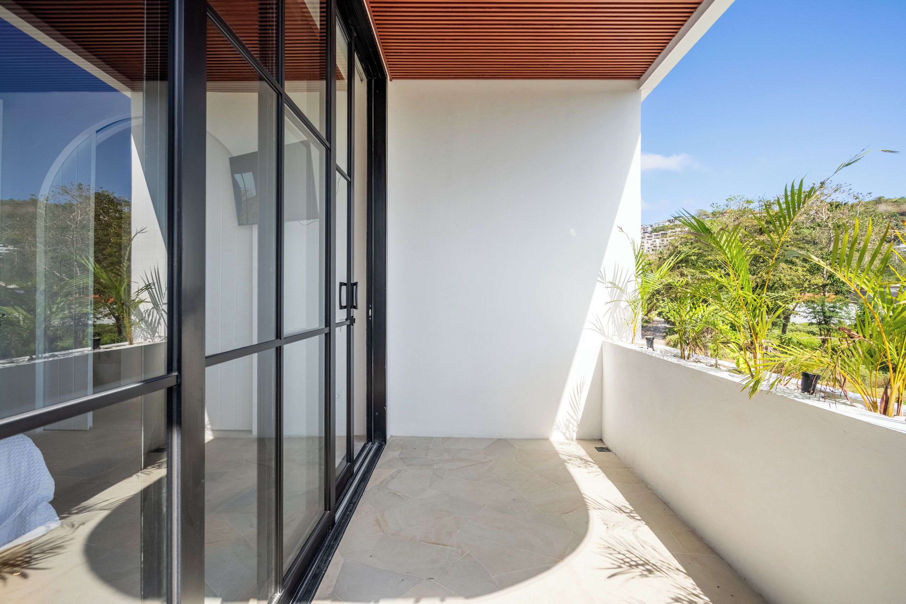 A modern balcony with glass doors, a white wall, and a view of greenery and buildings.