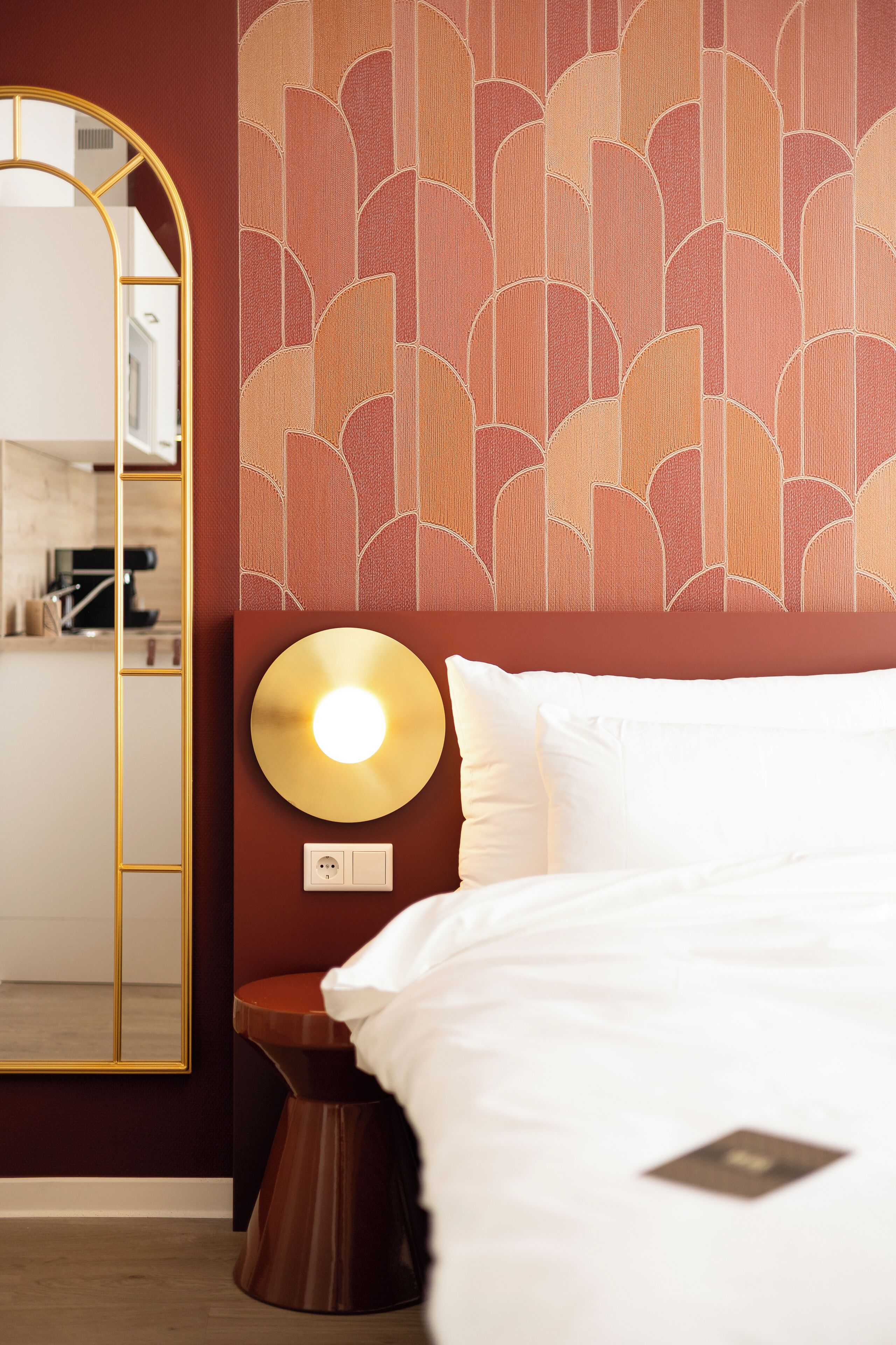 A hotel room with a bed, a bedside table with a lamp, a mirror, and a patterned wall.