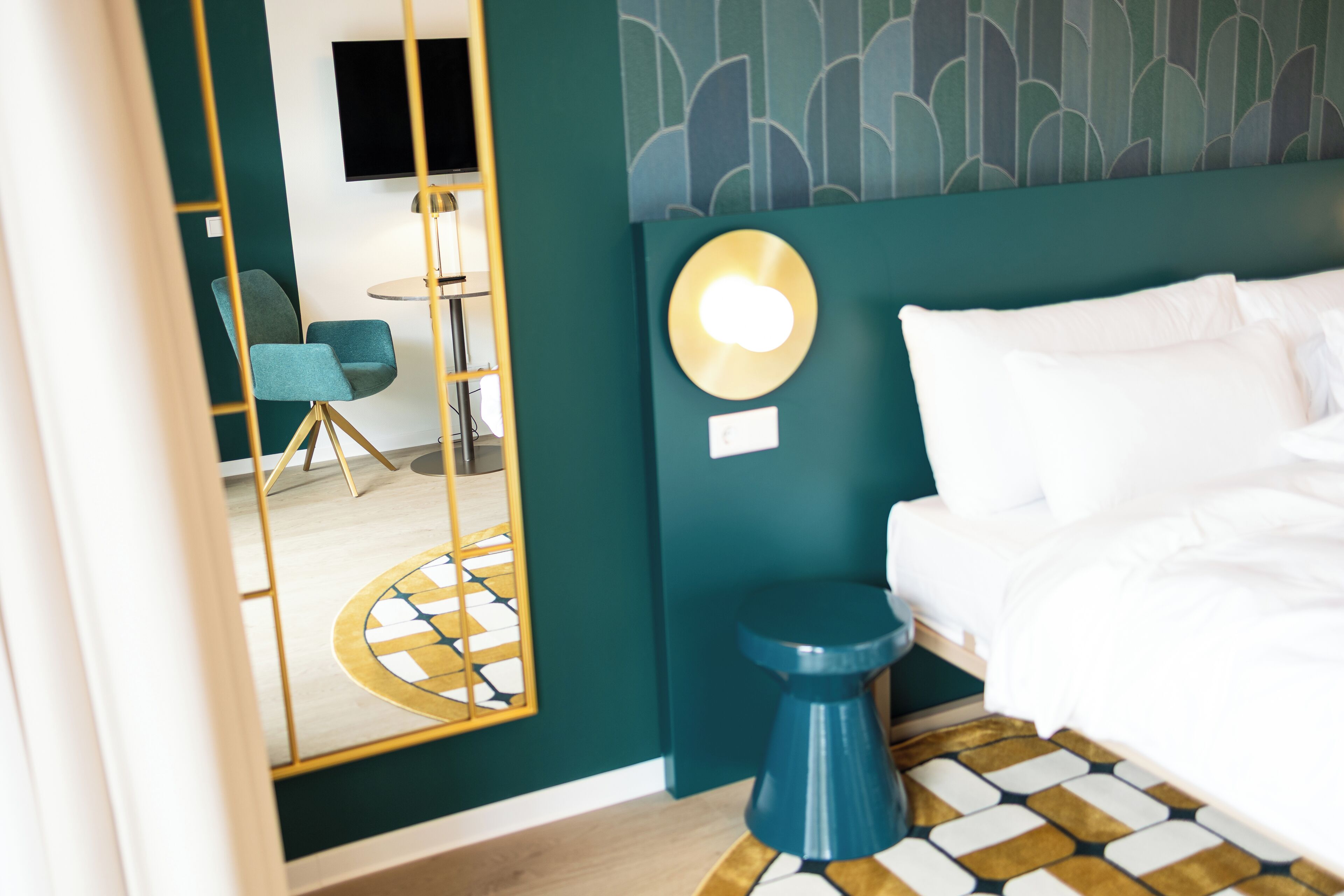 Habitación de hotel con cama, espejo redondo, taburete azul y pequeña mesa con lámpara.