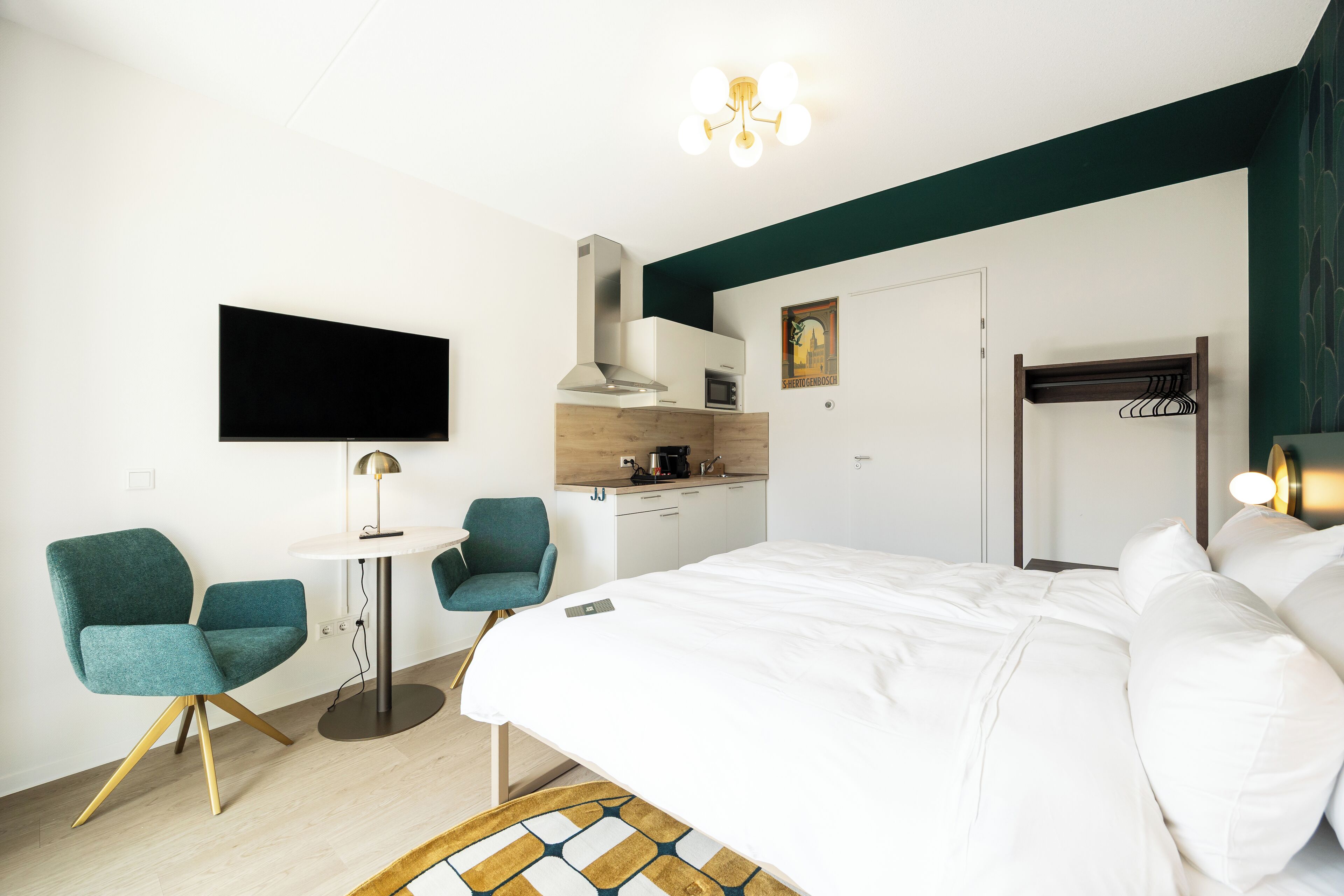 Habitación de hotel moderna con cama, dos sillas, una mesita, televisor, kitchenette y perchero.