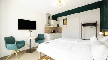 A modern hotel room with a bed, two chairs, a small table, a TV, a kitchenette, and a coat rack.