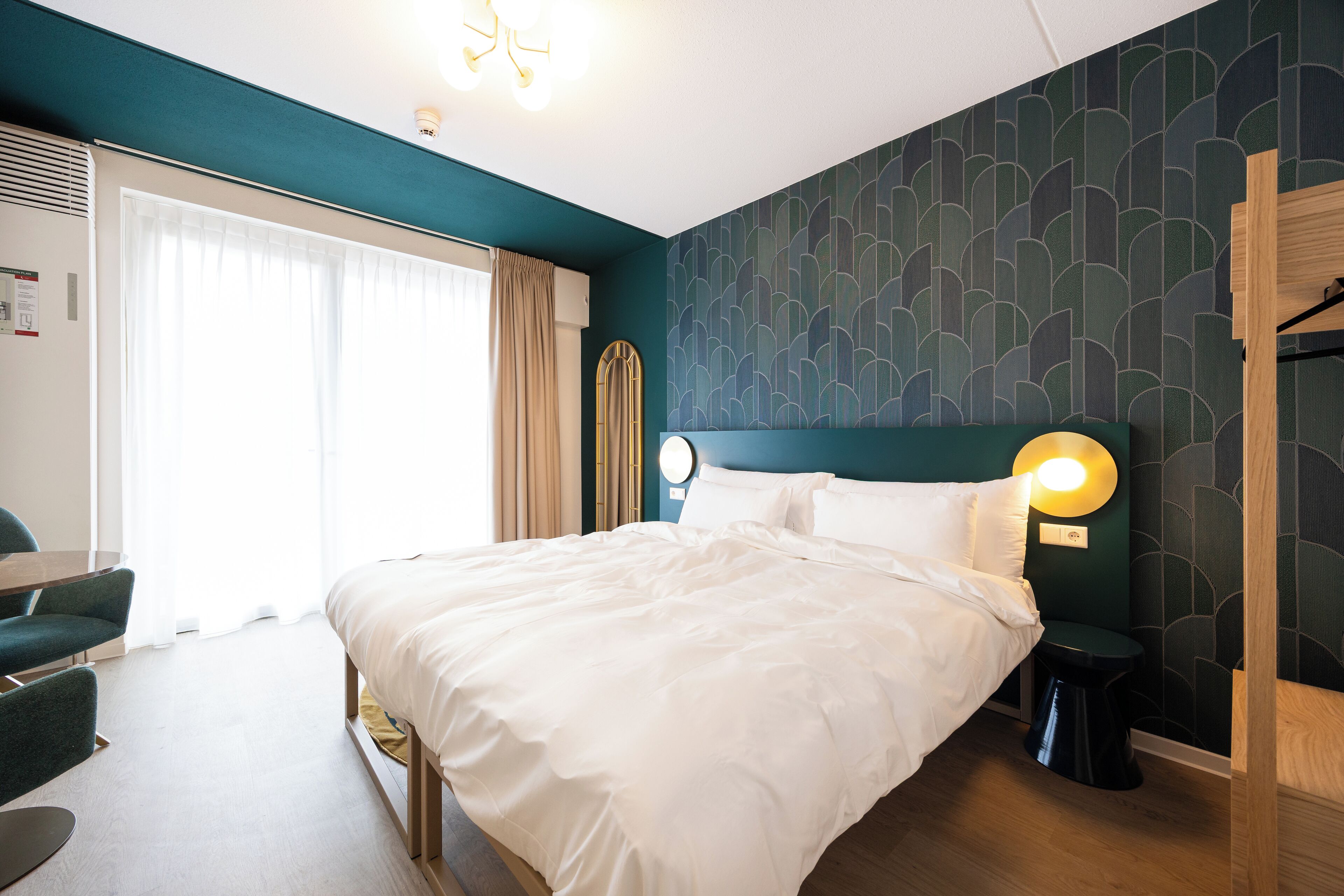 A hotel room with a large bed, a desk with a chair, a round mirror, and a bedside table with a lamp.