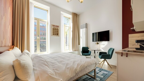 A hotel room with a large bed, a desk with a chair, a TV, and a view of buildings outside. - Duke Studio Hotel ('s-Hertogenbosch)