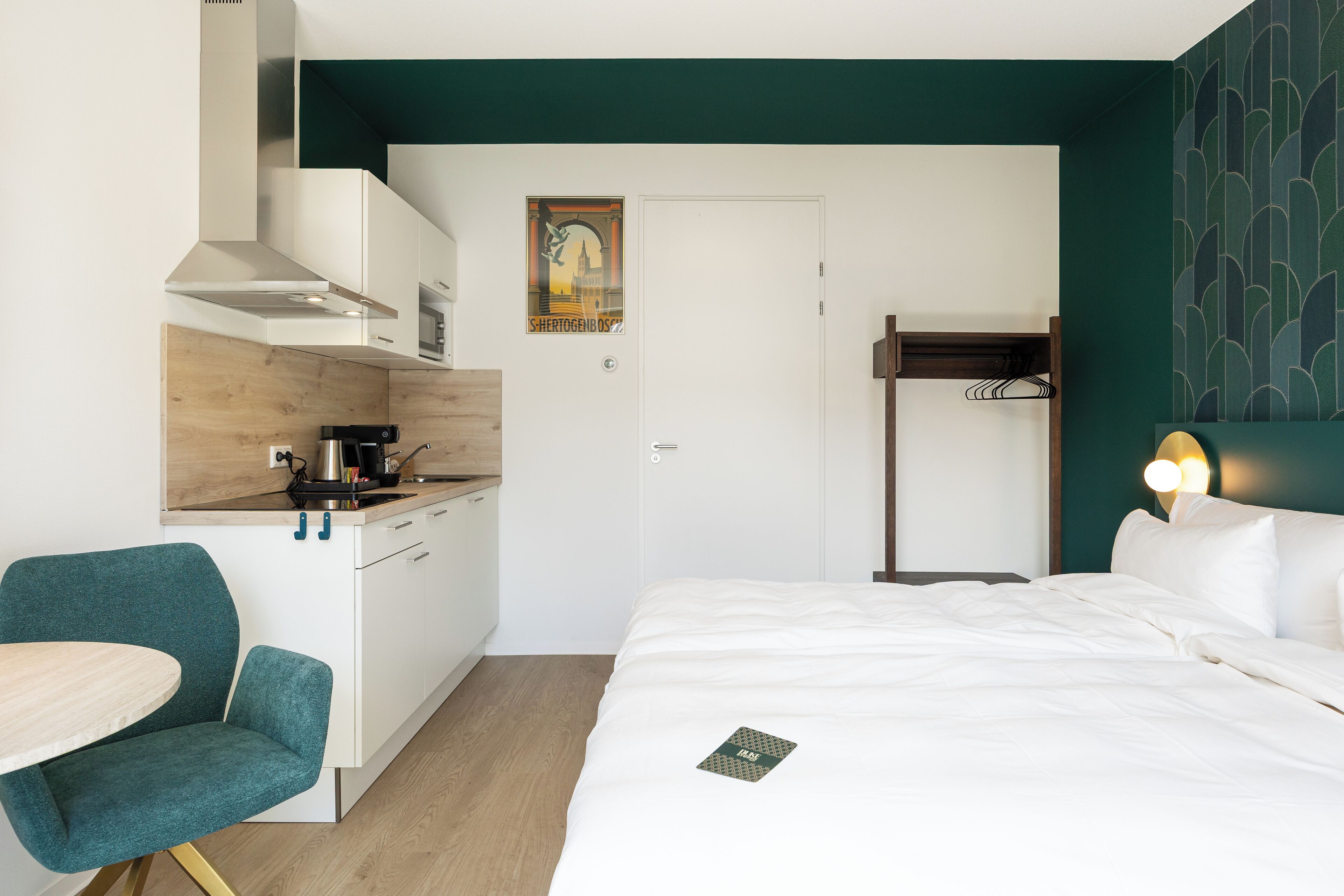 A compact hotel room with a kitchenette, a bed with white bedding, a teal chair, and a wall-mounted light fixture.