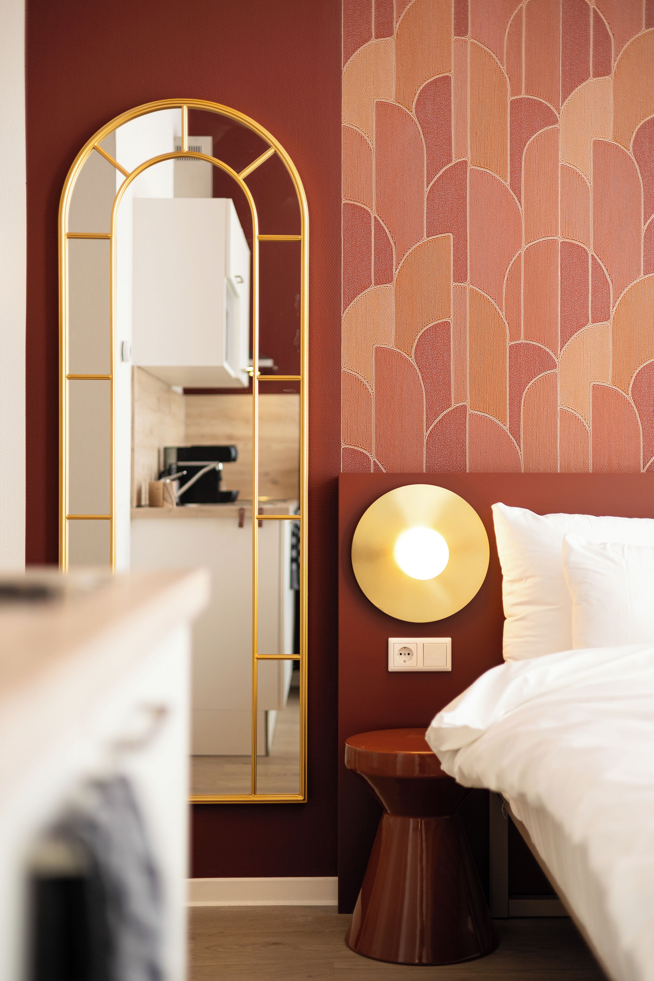 Habitación de hotel con cama, una lámpara redonda en la pared, espejo con marco dorado y pared con diseño.