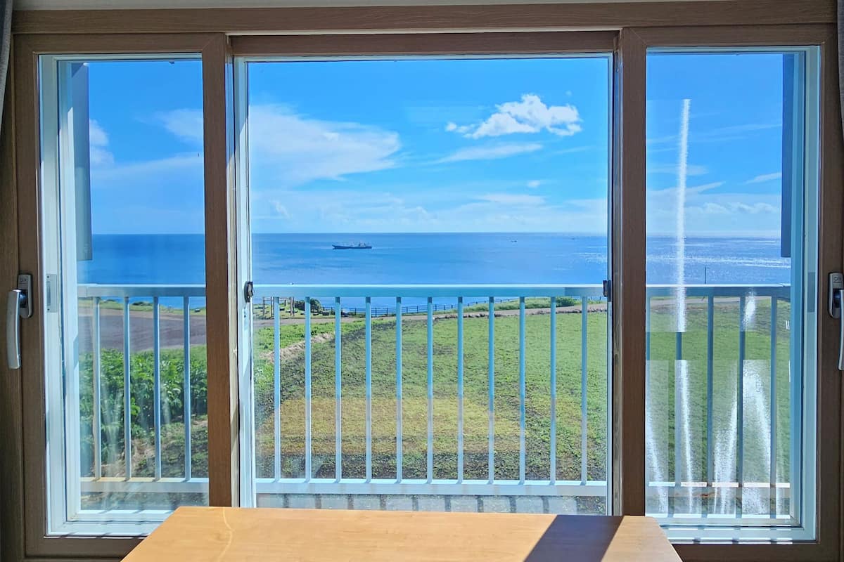 Une pièce dotée d’une grande fenêtre donnant sur un paysage côtier, avec vue sur l’océan et un navire.