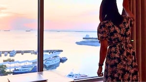 A woman in a patterned dress standing by a large window overlooking a marina with boats and a cruise ship. - Seaside Escape in Front of US Embassy (Manila)