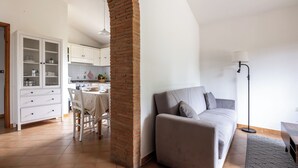 Una sala de estar moderna con un sofá, una mesa de comedor con sillas, una zona de cocina con armarios y un pilar de ladrillo.