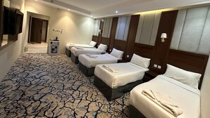 A hotel room with four beds arranged in a row, a small desk with a lamp, and a large mirror on the wall. - Manasik Al Bait Hotel Makkah (Makkah)