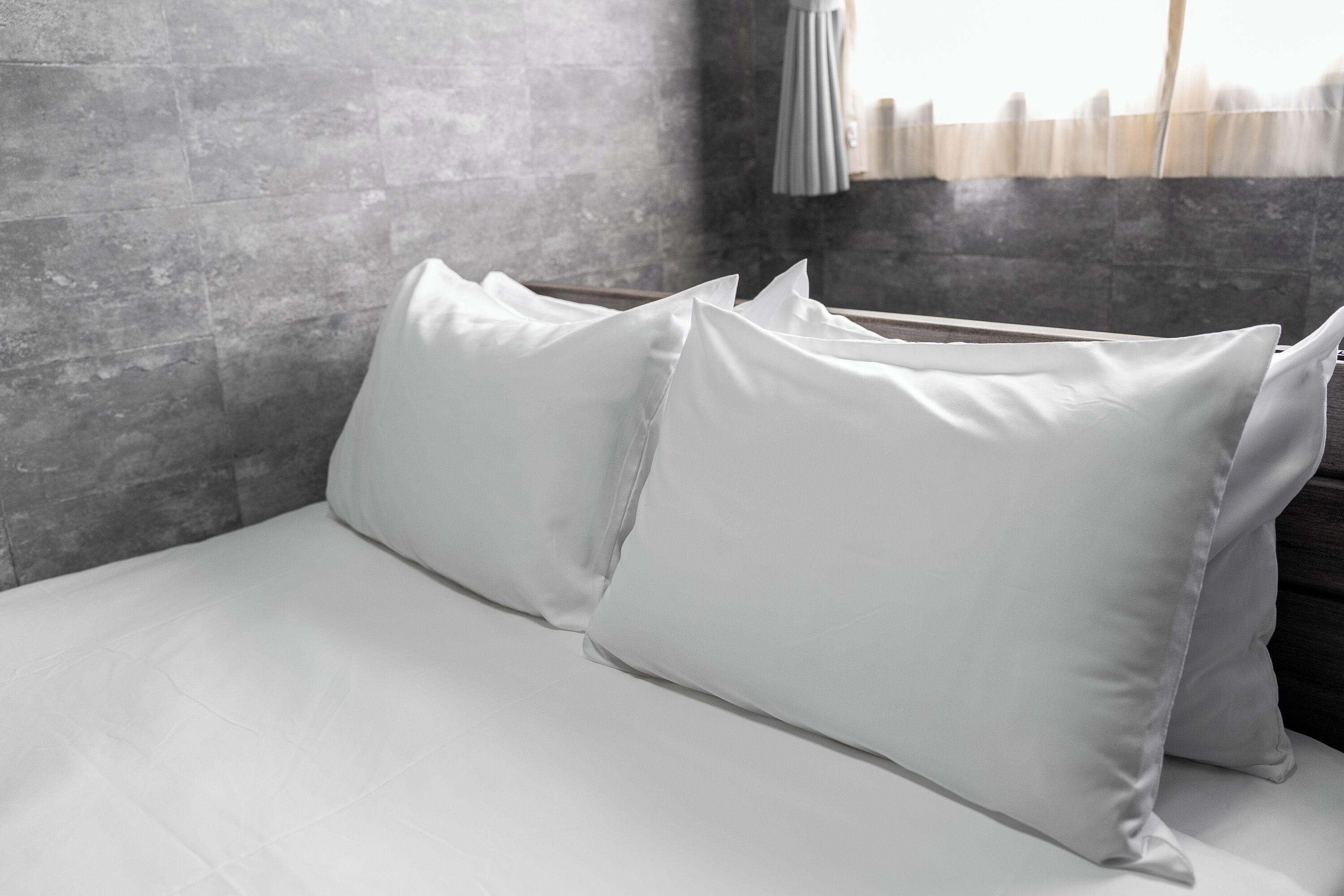 Una cama con sábanas y almohadas blancas en una habitación con pared de concreto y ventana con cortinas.