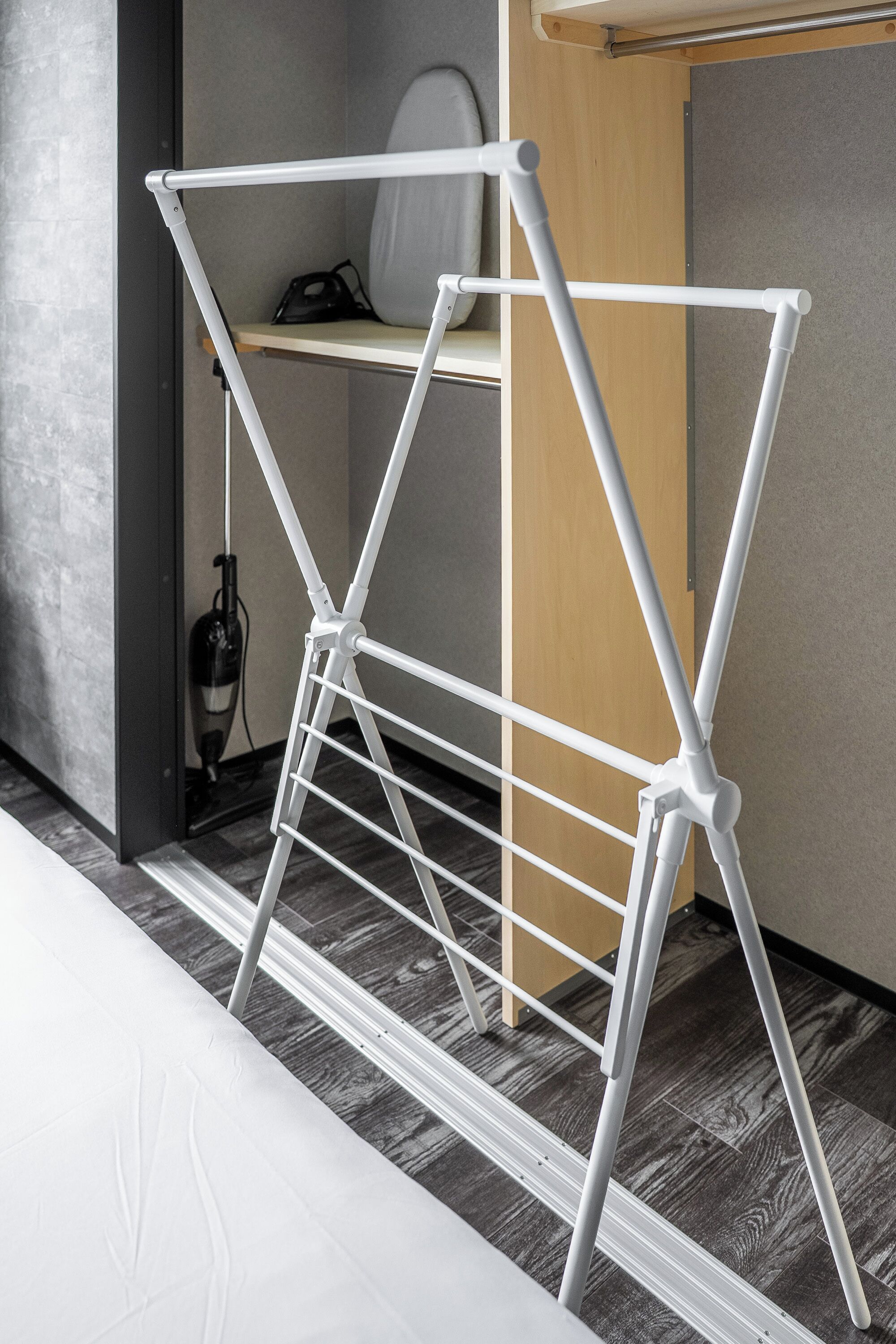 A white metal drying rack with a wooden shelf, positioned in a room with a grey wall and a wooden floor.