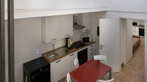 Cuina compacta amb rentatge, microones i fogó. Zona de menjador amb taula vermella i cadires blanques. La cuina té armaris blancs i una finestra petita sobre el rentatge.