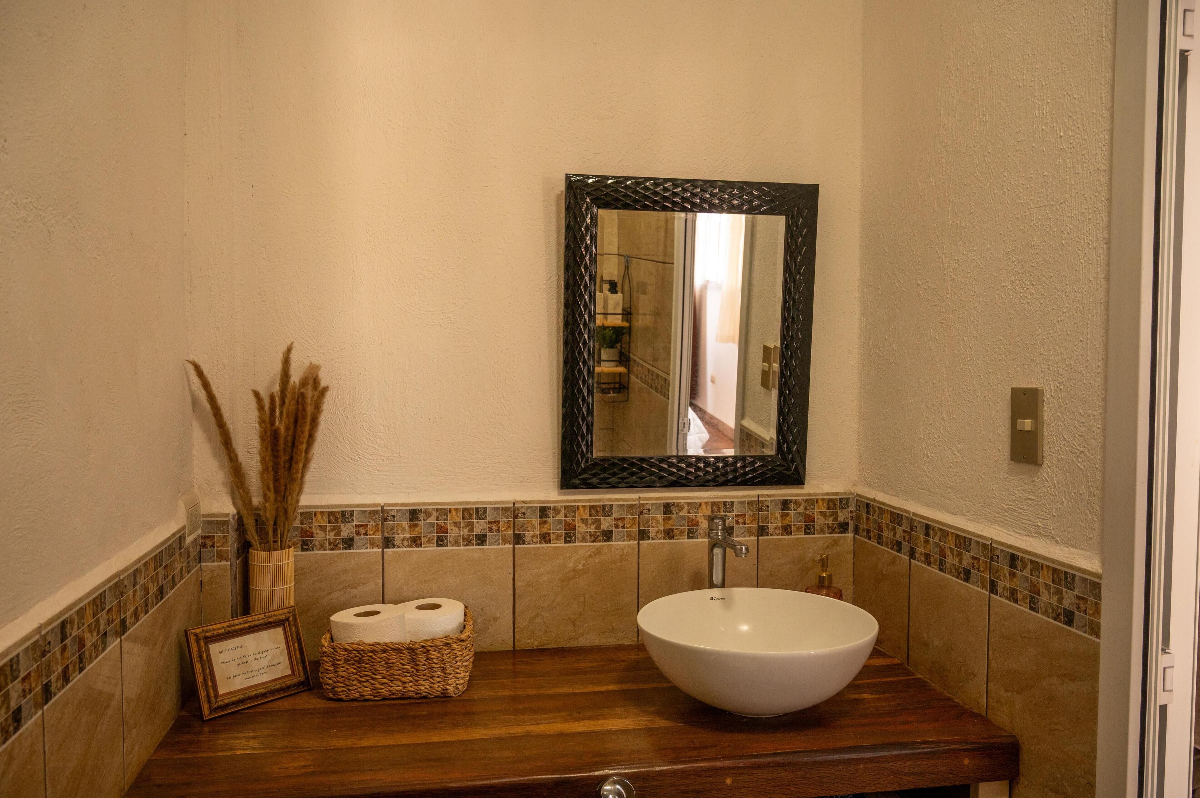 Une salle de bain avec un meuble-lavabo en bois, un lavabo blanc, un miroir avec un cadre noir et un pare-feu en carrelage.