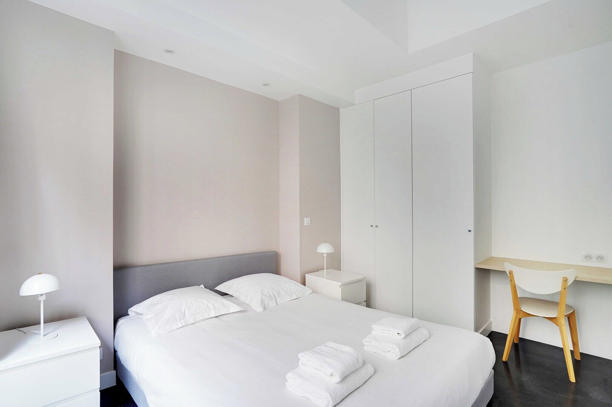 Una habitación de hotel moderna con una cama bien hecha, una mesita de noche con lámpara, un escritorio con silla y un armario.