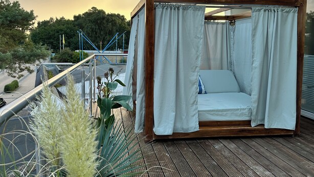 A wooden deck with a canopy bed, surrounded by plants and overlooking a body of water.