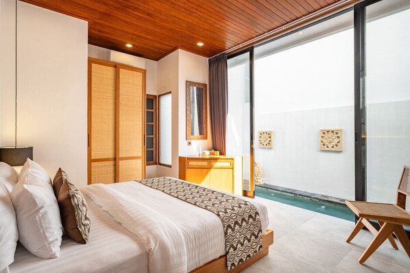 A hotel room with a bed, a wooden headboard, a bedside table, a mirror, a window, and a view of a swimming pool.