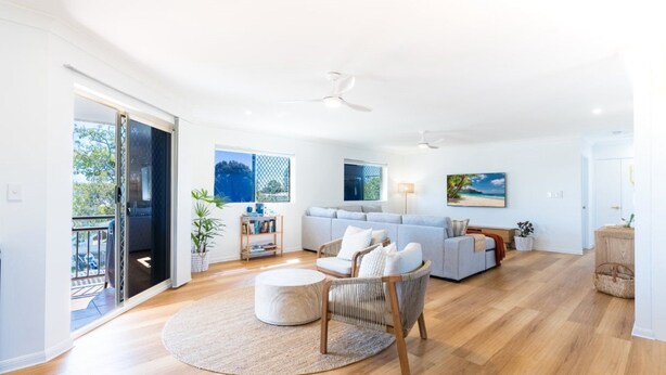 A spacious living room with a large sofa, a round coffee table, and a dining area with a wooden table and chairs. There is a balcony with a glass door and a view of the outdoors.