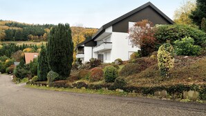A two-story residential building with a sloped roof, a balcony on the upper floor, and a well-maintained garden with various shrubs and trees.