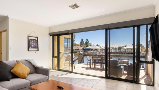 A modern living room with a grey sofa, a wooden coffee table, and a large sliding glass door leading to an outdoor patio with a view of a marina. - Canal apartment with Jetty (Halls Head)
