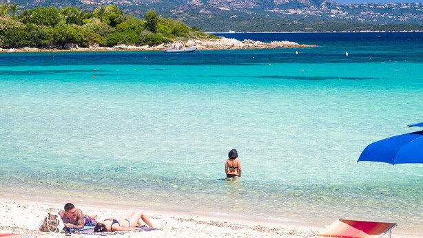Una playa con aguas turquesas cristalinas, una persona de pie en el agua y una cadena montañosa al fondo.