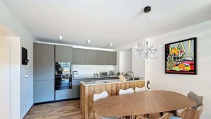 A modern kitchen with grey cabinets, a wooden dining table with six chairs, and a colorful abstract painting on the wall.